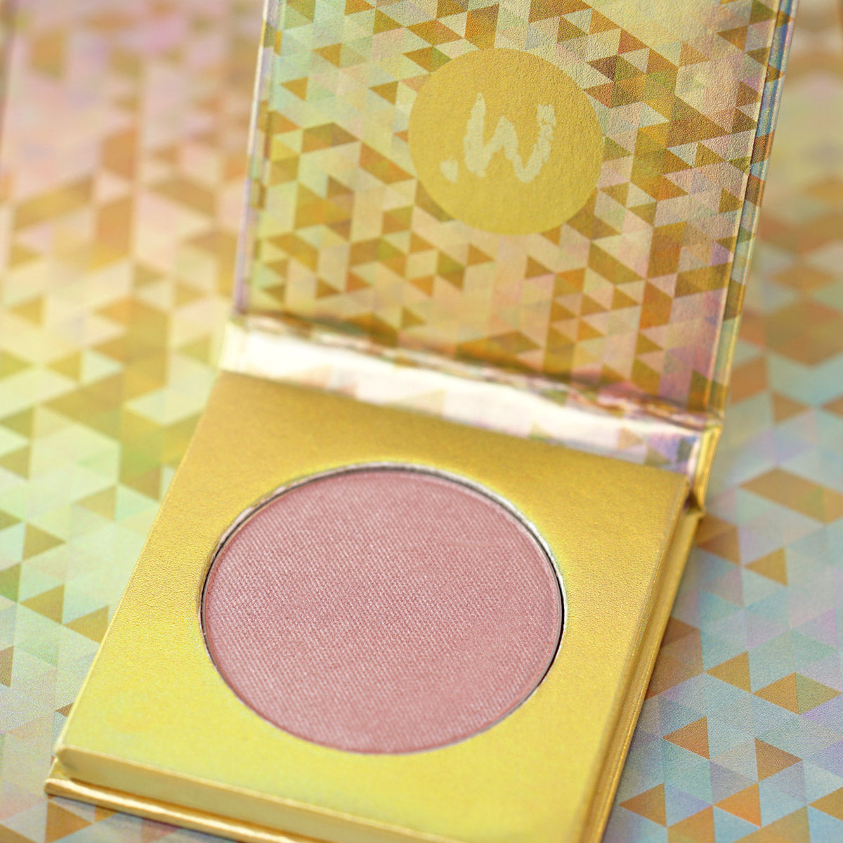 A compact pressed powder blush in a mauve plum shade, presented in a gold-colored case with a geometric pattern.